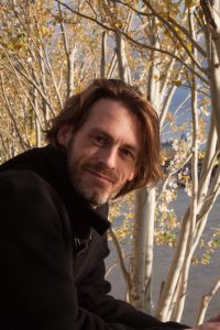 Dirk Primbs outdoors among bare birch trees in autumn, wearing a dark coat, hair loose.